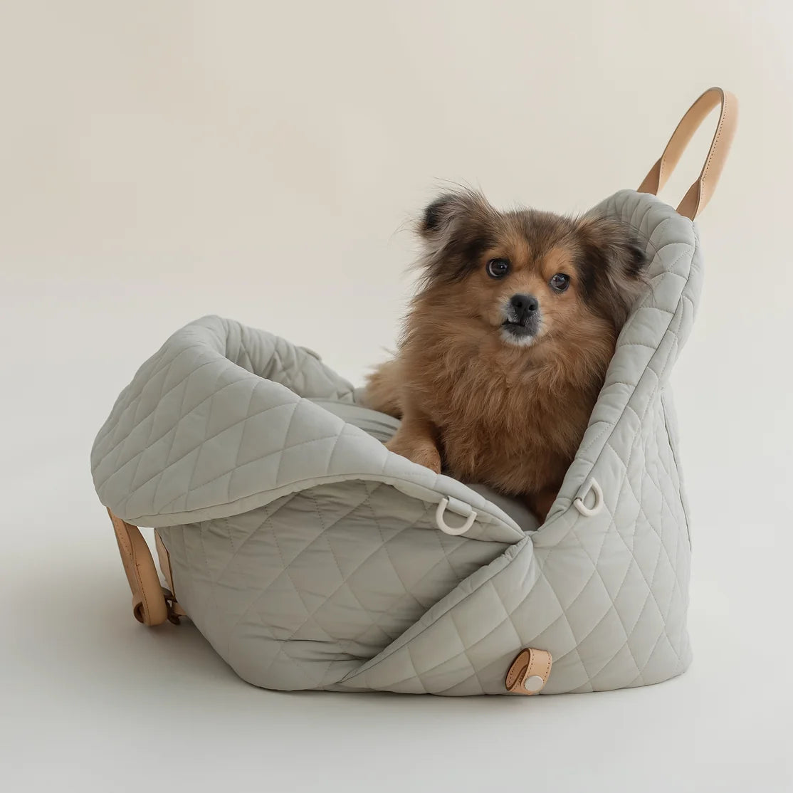 Ruby Max & Minette, A gray, quilted dog carrier or pet bed with leather accents and adjustable straps. A small, fluffy, brown dog with a black muzzle and ears is sitting inside a gray quilted pet bed.