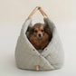 Ruby Max & Minette. A gray, quilted dog carrier or pet bed with leather accents and adjustable straps. A small, fluffy, brown dog with a black muzzle and ears is sitting inside a gray quilted pet bed, looking directly at the camera.