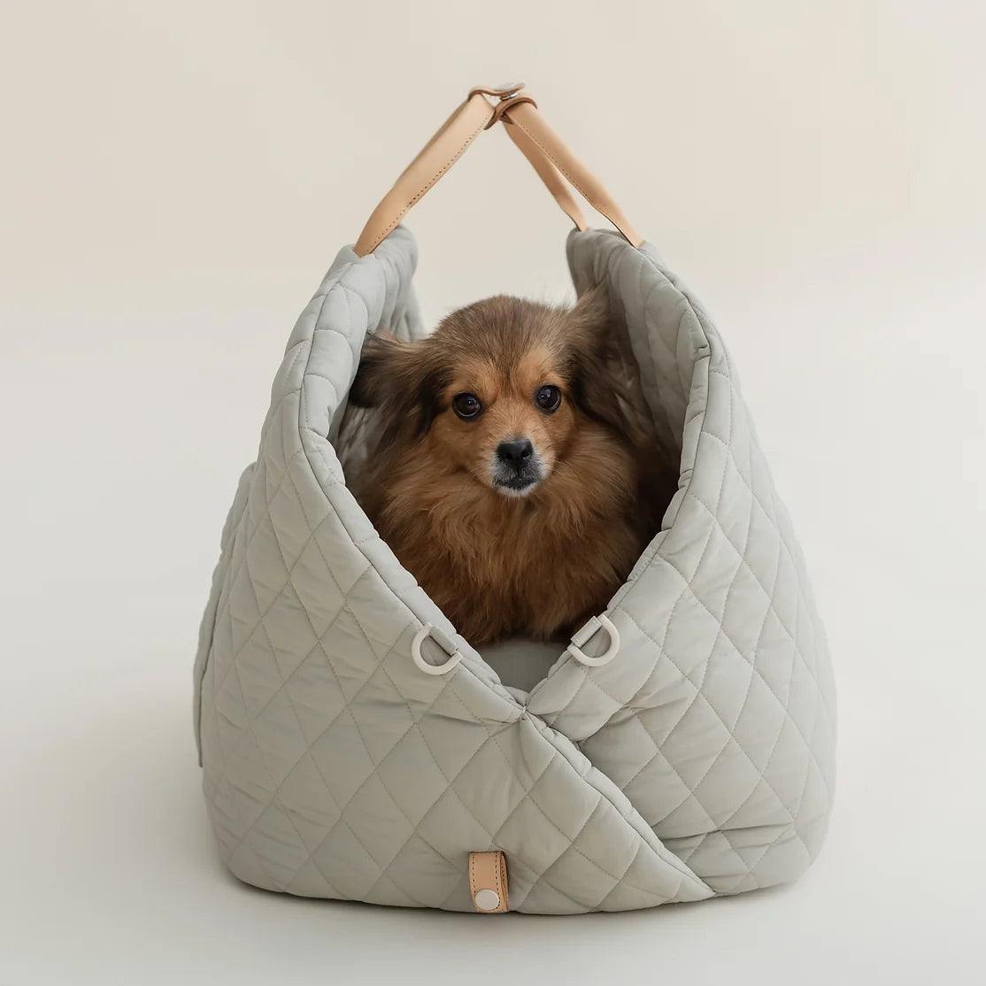 Ruby Max & Minette. A gray, quilted dog carrier or pet bed with leather accents and adjustable straps. A small, fluffy, brown dog with a black muzzle and ears is sitting inside a gray quilted pet bed, looking directly at the camera.