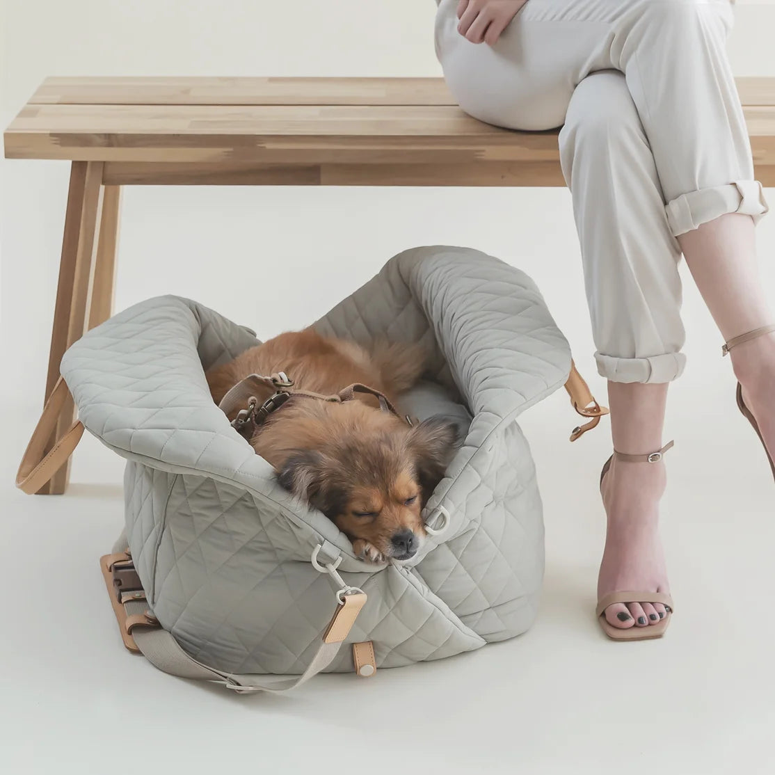 Ruby Max & Minette, A gray, quilted dog carrier or pet bed with leather accents and adjustable straps. A small, fluffy, brown dog with a black muzzle and ears is sitting inside a gray quilted pet bed