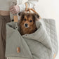 Ruby Max & Minette, A gray, quilted dog carrier or pet bed with leather accents and adjustable straps. A small, fluffy, brown dog with a black muzzle and ears is sitting inside a gray quilted pet bed, looking directly at the camera.