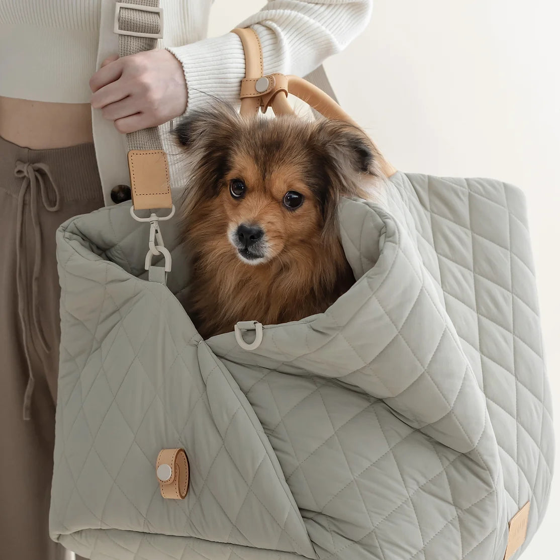 Ruby Max & Minette, A gray, quilted dog carrier or pet bed with leather accents and adjustable straps. A small, fluffy, brown dog with a black muzzle and ears is sitting inside a gray quilted pet bed, looking directly at the camera.