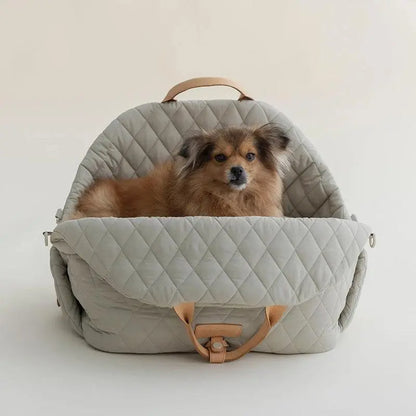 A gray, quilted dog carrier or pet bed with leather accents and adjustable straps. A small, fluffy, brown dog with a black muzzle and ears is sitting inside a gray quilted pet bed, looking directly at the camera.